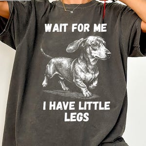 Op de afbeelding: Donkergrijs t-shirt met een gedetailleerde illustratie van een teckel. De tekst "WAIT FOR ME I HAVE LITTLE LEGS" is in het wit gedrukt. Het shirt heeft een relaxte pasvorm en een vintage esthetiek.