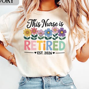 May include: Ivory-colored t-shirt with the text "This Nurse is Retired Est. 2026" in black, pink, yellow, blue, and green. The design includes colorful flowers above the word "Retired". A casual, comfortable tee.