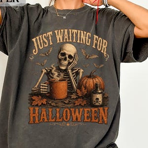 May include: A charcoal gray t-shirt featuring a graphic of a skeleton resting its chin on one hand while holding a mug. The design includes a pumpkin, autumn leaves, bats, a small bottle with a skull label, and the text JUST WAITING FOR HALLOWEEN.
