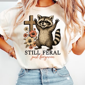 May include: Ivory-colored t-shirt featuring a raccoon illustration with a cross and flowers. The raccoon has its arms raised, and the text reads "STILL FERAL just forgiven". The shirt is a casual wear item.