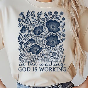 May include: Cream-colored t-shirt featuring a navy blue floral design with the text "in the waiting GOD IS WORKING". The graphic includes stylized flowers and foliage, creating a botanical theme.