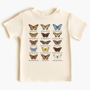 May include: Cream-colored t-shirt featuring a colorful butterfly chart. The chart includes illustrations of various butterfly species with their names, such as Monarch, Swallowtail, and Blue Morpho. The shirt is made of soft fabric.