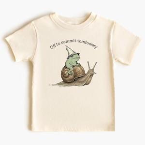 Op de afbeelding: Crèmekleurig T-shirt met een grillige illustratie van een groene kikker met een tovenaars hoed op een slak. De tekst "Off to commit tomfoolery" staat boven de afbeelding. Het shirt is gemaakt van zacht materiaal.