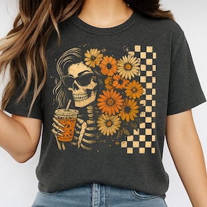 May include: Dark gray t-shirt featuring a skeleton with sunglasses, holding a drink, and surrounded by orange and yellow flowers. A checkered pattern is on the right side of the design. The shirt is a casual, comfortable style.