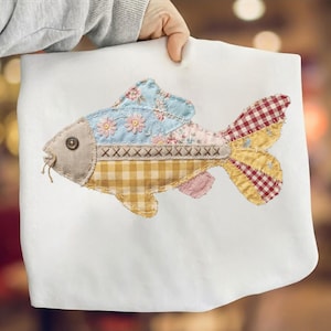 Può includere: Applicazione decorativa a forma di pesce su sfondo bianco. Il pesce è realizzato con vari tessuti fantasia, tra cui floreale, gingham e scozzese. Ha un occhio marrone e una bocca cucita. Le pinne sono un mix di tessuti gialli, rossi e blu.