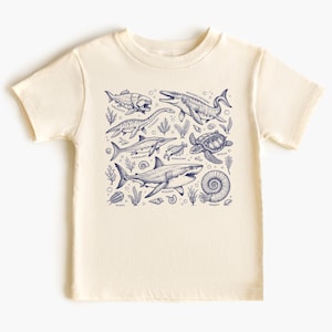 Könnte beinhalten: Cremefarbenes T-Shirt mit einer marineblauen Illustration verschiedener prähistorischer Meeresbewohner, darunter Haie und Schildkröten. Das Design umfasst wissenschaftliche Namen und dekorative Seetang-Elemente.