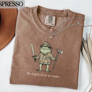 Op de afbeelding: Espresso-kleurig T-shirt met een cartoon kikker brandweerman met een zwaard en een bijl. De kikker draagt een brandweerhelm en jas. De tekst "We fight fire at dawn" is eronder gedrukt. Een zilveren ketting ligt over het shirt.