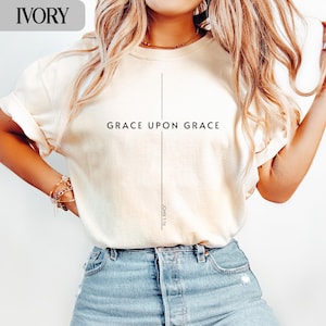 May include: Ivory-colored t-shirt with the text "GRACE UPON GRACE" in black, centered on the front. A vertical line intersects the text, forming a cross. The text "JOHN 1:16" is printed vertically below the cross.