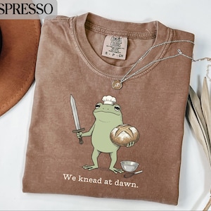 May include: Espresso-colored t-shirt featuring a cartoon frog chef holding a loaf of bread and a sword. The frog wears a chef's hat and the text "We knead at dawn" is printed below. A silver necklace is draped across the shirt.
