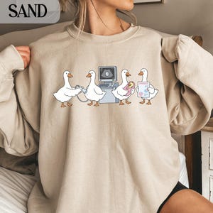 Op de afbeelding: Zandkleurige sweatshirt met een humoristische illustratie van ganzen in een medische setting. De ganzen worden afgebeeld met een echografieapparaat, een baby vasthoudend en een medisch dossier. Het woord "SAND" is zichtbaar in de linkerbovenhoek.