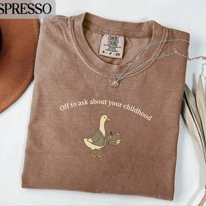 Peut inclure: T-shirt couleur expresso avec l'inscription "Off to ask about your childhood" et un dessin de canard en costume, tenant un presse-papiers. Le t-shirt a un col rond et une étiquette Comfort Colors. Un collier argenté avec un pendentif est posé sur le t-shirt.
