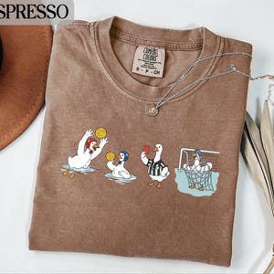 以下が含まれることがあります： エスプレッソ色のクルーネックTシャツ。水球とサッカーをするアヒルの風変わりなグラフィックが特徴です。アヒルはヘルメットと審判のユニフォームを着ています。ペンダント付きのシルバーネックレスも確認できます。