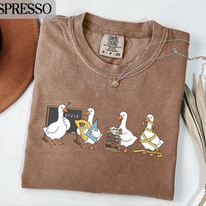 May include: Espresso-colored t-shirt featuring a whimsical design of four cartoon geese. Each goose is depicted with school supplies, including a chalkboard, protractor, abacus, and measuring tape. The shirt has a crew neck and a Comfort Colors tag.
