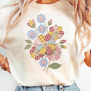 Op de afbeelding: Crèmekleurig T-shirt met een kleurrijk bloemenontwerp. De bloemen zijn gemaakt van patchworkstof in roze, gele, blauwe en rood geruite tinten. Het ontwerp is gecentreerd op het shirt, met groene bladeren en stelen.