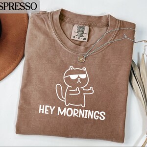 May include: Espresso-colored t-shirt with a white graphic of a cat wearing sunglasses and giving the middle finger. The text "HEY MORNINGS" is below the cat. A silver necklace with a small pendant is on the shirt. The shirt is on a white surface.