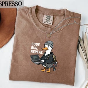 May include: Espresso-colored t-shirt with a graphic of a duck wearing a beanie and backpack, holding a laptop, keyboard, and mouse. The text "CODE. BUG. REPEAT." is printed on the shirt. A silver necklace is draped across the shirt.