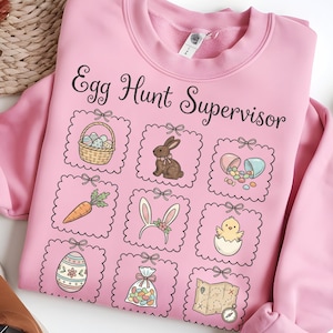 May include: Pink sweatshirt with the text "Egg Hunt Supervisor" and Easter-themed graphics. The design includes a basket of eggs, a bunny, a carrot, bunny ears, a chick, an egg, a bag of jelly beans, and a map.