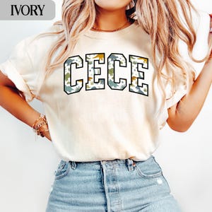 May include: Ivory-colored sweatshirt with the name "CECE" in large, block letters. The letters are filled with a pattern of green, blue, and yellow dinosaurs. The sweatshirt is a casual style, suitable for everyday wear.