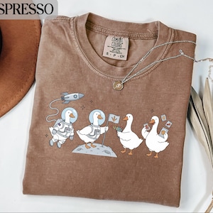 以下が含まれることがあります： エスプレッソ色のTシャツ。宇宙服を着た漫画のガチョウの気まぐれなデザインが特徴です。1羽のガチョウは宇宙服を着ており、別のガチョウは旗を立て、他の2羽はアイテムを持っています。シャツはクルーネックで、Comfort Colorsのラベルが付いています。