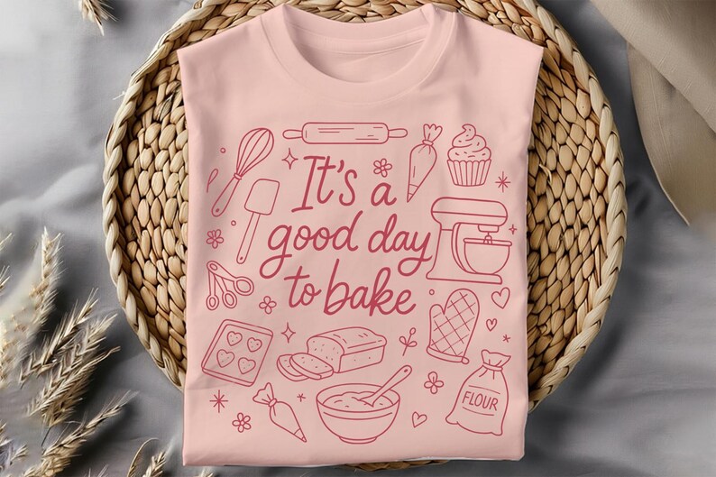 May include: A light pink t-shirt with a baking-themed design. The design includes a whisk, rolling pin, mixer, and the text "It's a good day to bake" in a handwritten style. The shirt is displayed on a woven basket.
