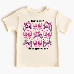May include: Cream-colored t-shirt featuring a design with pink bows, video game controllers, and hearts. The text "Girls like video games too" is printed in black. The design is centered on the front of the shirt.