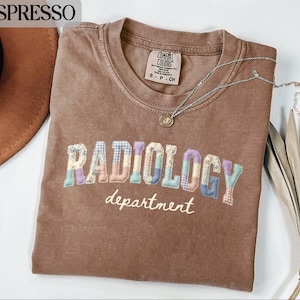 May include: Espresso-coloured t-shirt with the word "RADIOLOGY" in patchwork letters and "department" in script. A silver necklace with a pendant rests on the shirt. The shirt is a Comfort Colors brand.