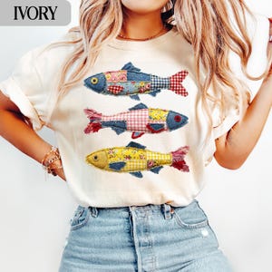 May include: Ivory t-shirt with three fish designs. Each fish is made from patchwork denim, floral, and gingham fabrics in blue, red, and yellow. The shirt has a relaxed fit. The word "IVORY" is printed at the top.