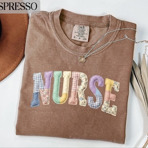 Pode incluir: Camiseta cor café com a palavra "NURSE" aplicada na frente em tecido patchwork. As letras são em várias cores e padrões. Um colar de prata com um pequeno pingente repousa sobre a camiseta. A camiseta é da marca Comfort Colors.