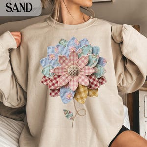 Könnte beinhalten: Ein sandfarbenes Sweatshirt mit einem großen, applizierten Blumenmuster. Die Blume hat geschichtete Blütenblätter aus verschiedenen gemusterten Stoffen, darunter Vichy, Blumenmuster und unifarbene Farben. Stiel und Blätter sind ebenfalls appliziert.
