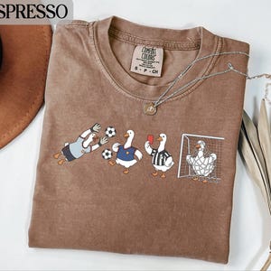 May include: Espresso-colored t-shirt featuring a soccer-themed graphic with cartoon geese. One goose is a goalie, others are players, and one is a referee. The shirt has a crew neck and short sleeves. A silver necklace with a pendant is on the shirt.