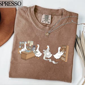 Op de afbeelding: Espresso-kleurig T-shirt met een cartoon van ganzen die in een apotheek werken. De ganzen zijn te zien achter een toonbank, bij een kassa en schappen aan het vullen. Het shirt heeft een ronde hals en een Comfort Colors-label.