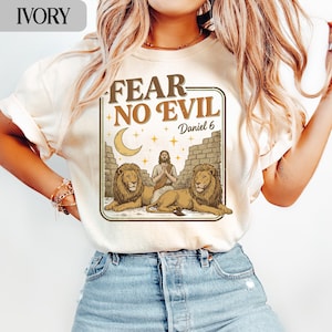May include: Ivory-colored t-shirt with a graphic design featuring the text "FEAR NO EVIL" and "Daniel 6". The design includes an illustration of a figure with lions, a crescent moon, and stars.