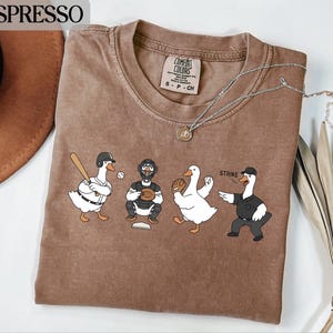 Könnte beinhalten: Espressofarbenes T-Shirt mit einem Baseball-Motiv mit Cartoon-Gänsen. Eine Gans hält einen Schläger, eine andere einen Baseball, eine dritte einen Handschuh und die letzte ist ein Schiedsrichter. Das Wort "STRIKE" steht über der Schiedsrichter-Gans.