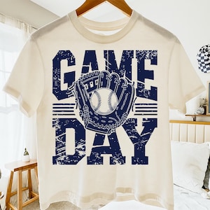 以下が含まれることがあります： クリーム色のTシャツにネイビーブルーのグラフィック。グラフィックは、野球のグローブと野球ボールを中央に配置し、かすれたフォントで「GAME DAY」という言葉を表示しています。木製のハンガーにTシャツが掛かっています。