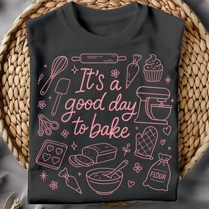 May include: A dark gray t-shirt with a pink outline design featuring baking tools and the phrase "It's a good day to bake." The design includes a whisk, rolling pin, mixer, and other baking-related graphics. The shirt is displayed on a woven basket.