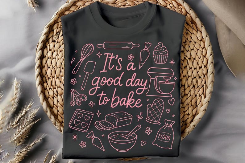 May include: A dark gray t-shirt with a pink outline design featuring baking tools and the phrase "It's a good day to bake." The design includes a whisk, rolling pin, mixer, and other baking-related graphics. The shirt is displayed on a woven basket.