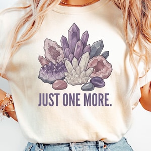 Peut inclure: T-shirt crème avec un motif de cristaux violets, blancs et gris, et le texte "JUST ONE MORE." Les cristaux sont disposés avec de petites pierres en bas. Un haut décontracté.