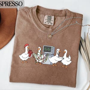 Könnte beinhalten: Espressofarbenes T-Shirt mit einem skurrilen Design von Cartoon-Gänsen in medizinischen Rollen. Eine Gans hält ein Herz, eine andere ist mit Drähten verbunden, und zwei andere benutzen ein Stethoskop und geben einen Daumen hoch. Das Shirt hat einen Rundhalsausschnitt und kurze Ärmel.