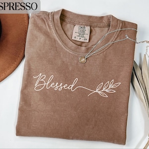 Könnte beinhalten: Espressofarbenes T-Shirt mit dem Wort "Blessed" in weißer Schrift und einem Blattdesign. Das Shirt hat einen Rundhalsausschnitt und kurze Ärmel. Eine silberne Halskette mit einem kleinen Anhänger liegt über dem Shirt.