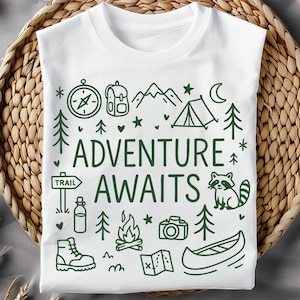 May include: White t-shirt with a green graphic design featuring outdoor adventure icons such as a compass, tent, and canoe. The words "ADVENTURE AWAITS" are displayed. The shirt is folded on a woven basket.
