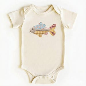 Op de afbeelding: Crème babybody met een kleurrijke patchwork vis applicatie. De vis heeft blauwe bloemen, bruine ruitjes en rode ruitpatronen. De body heeft korte mouwen en drukknoopsluitingen.