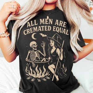 以下が含まれることがあります： 「ALL MEN ARE CREMATED EQUAL」の文字と、骸骨と魔女のグラフィックが描かれた黒いTシャツ。骸骨は大釜の中にいて、魔女は杖を持っています。コウモリと三日月もデザインに含まれています。