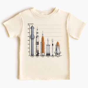 May include: A cream-coloured t-shirt displaying a graphic of several rockets with height measurements in feet. The rockets are in shades of grey, orange, and white. The text "FEET (Height):" is at the top.