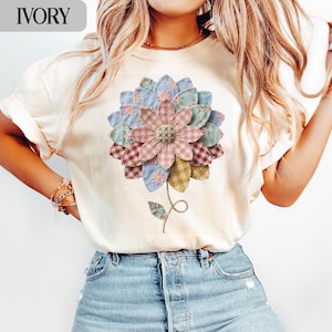 May include: Cream-coloured t-shirt with a large floral design made from patchwork fabric. The flower features pink, blue, green, and yellow patches. The word "IVORY" is printed at the top left of the t-shirt.