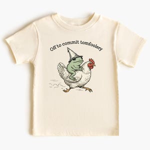 以下が含まれることがあります： クリーム色のTシャツ。奇抜なイラストが描かれています。緑色のカエルが魔法使いの帽子をかぶり、白い鶏に乗っています。「Off to commit tomfoolery」というテキストが画像の上にあります。このシャツは柔らかい素材で作られています。