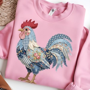 May include: A pale pink sweatshirt with an appliqué of a rooster. The rooster design uses patterned fabrics, including floral, polka dot, and gingham, in blue, red, and white. The rooster's comb is red, and its beak is yellow.