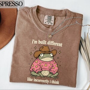 May include: An espresso-colored t-shirt featuring a graphic of a green frog wearing a brown cowboy hat and a pink sweater with yellow stars. The text reads "i'm built different" above the frog and "like incorrectly i think" below the frog.