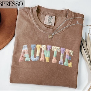May include: Espresso-colored t-shirt with the word "AUNTIE" in colorful patchwork letters. The shirt has a crew neck and a small tag on the upper back. A silver necklace with a pendant is draped across the shirt. A brown hat is visible in the upper left corner.
