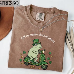 以下が含まれることがあります： 「Off to cause shenanigans」のテキストが入ったエスプレッソカラーのTシャツ。緑のレプラコーンハットをかぶったカエルの漫画が、緑のスクーターに乗っています。金貨とクローバーがカエルとスクーターの周りに散らばっています。