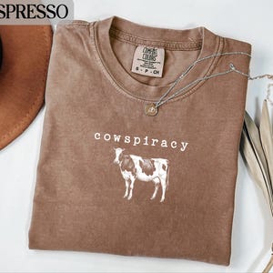 Op de afbeelding: Espresso-kleurig T-shirt met het woord "cowspiracy" en een koe-afbeelding. Het shirt heeft een ronde hals en korte mouwen. Een zilveren ketting met een hanger is over het shirt gedrapeerd. Een bruine hoed en gedroogde bladeren zijn op de achtergrond.